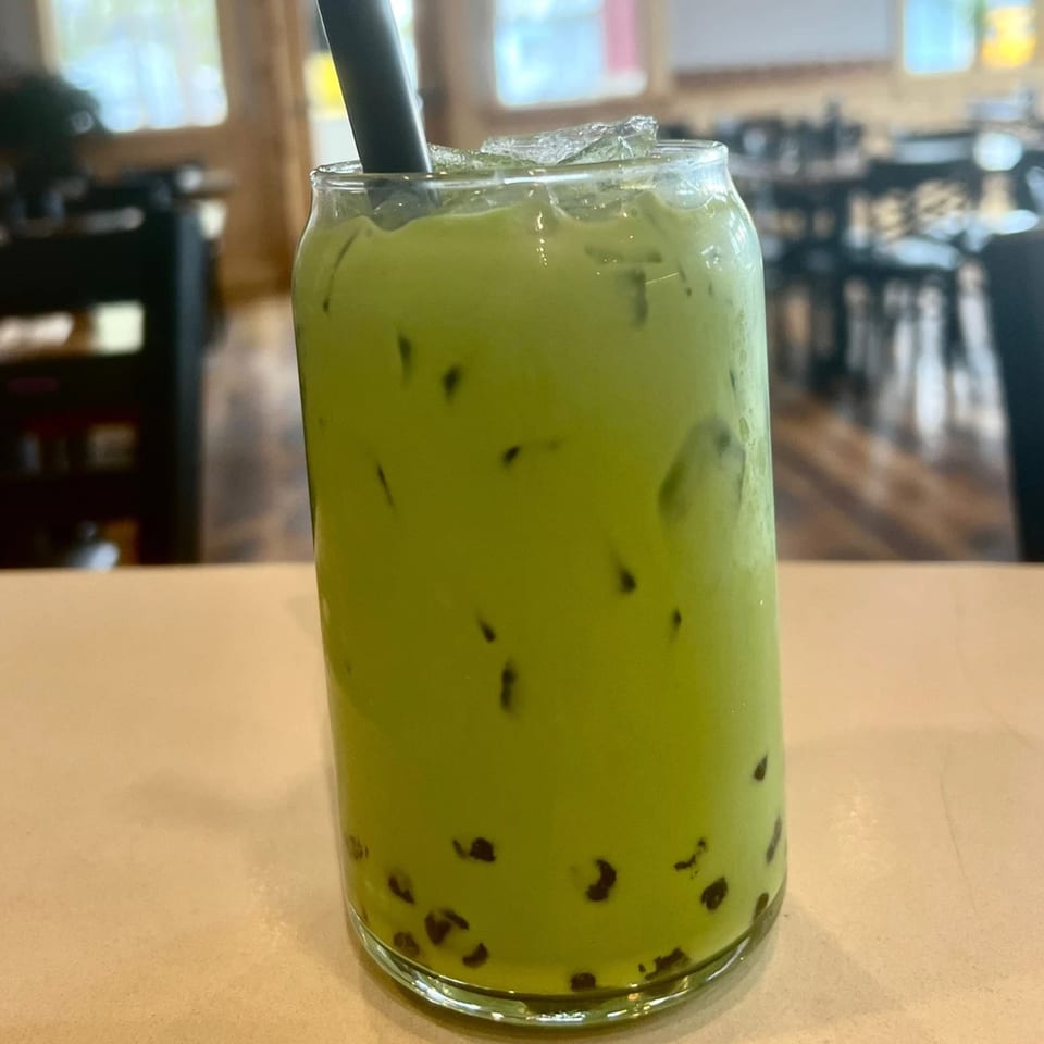 A tall glass of green Thai iced tea with boba and ice, with a black straw.